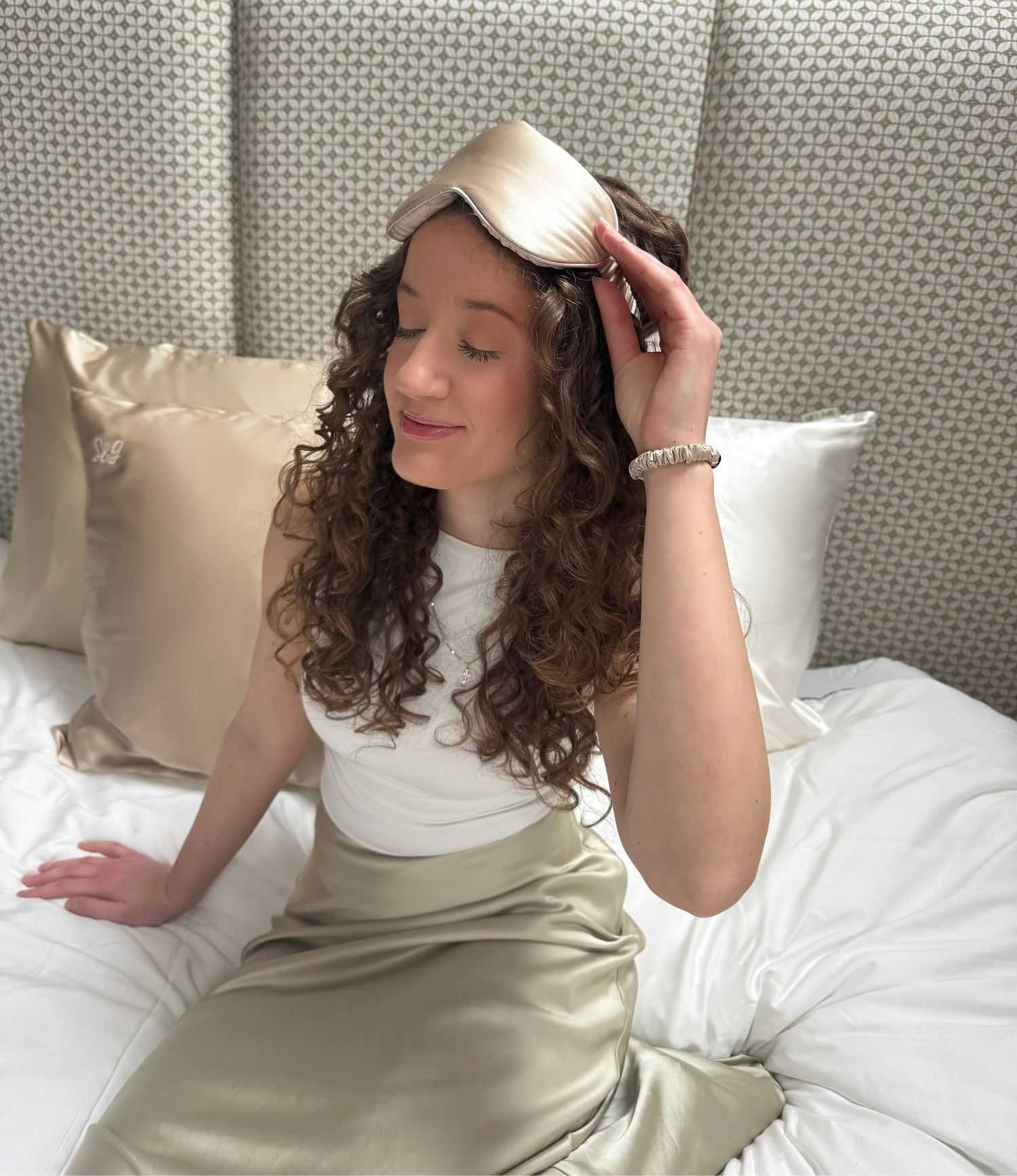 Woman sitting on a bed wearing a gold Mulberry Silk sleepmask and hair ribbon, with a neutral-colored headboard and Mulberry Silk pillows in the background.